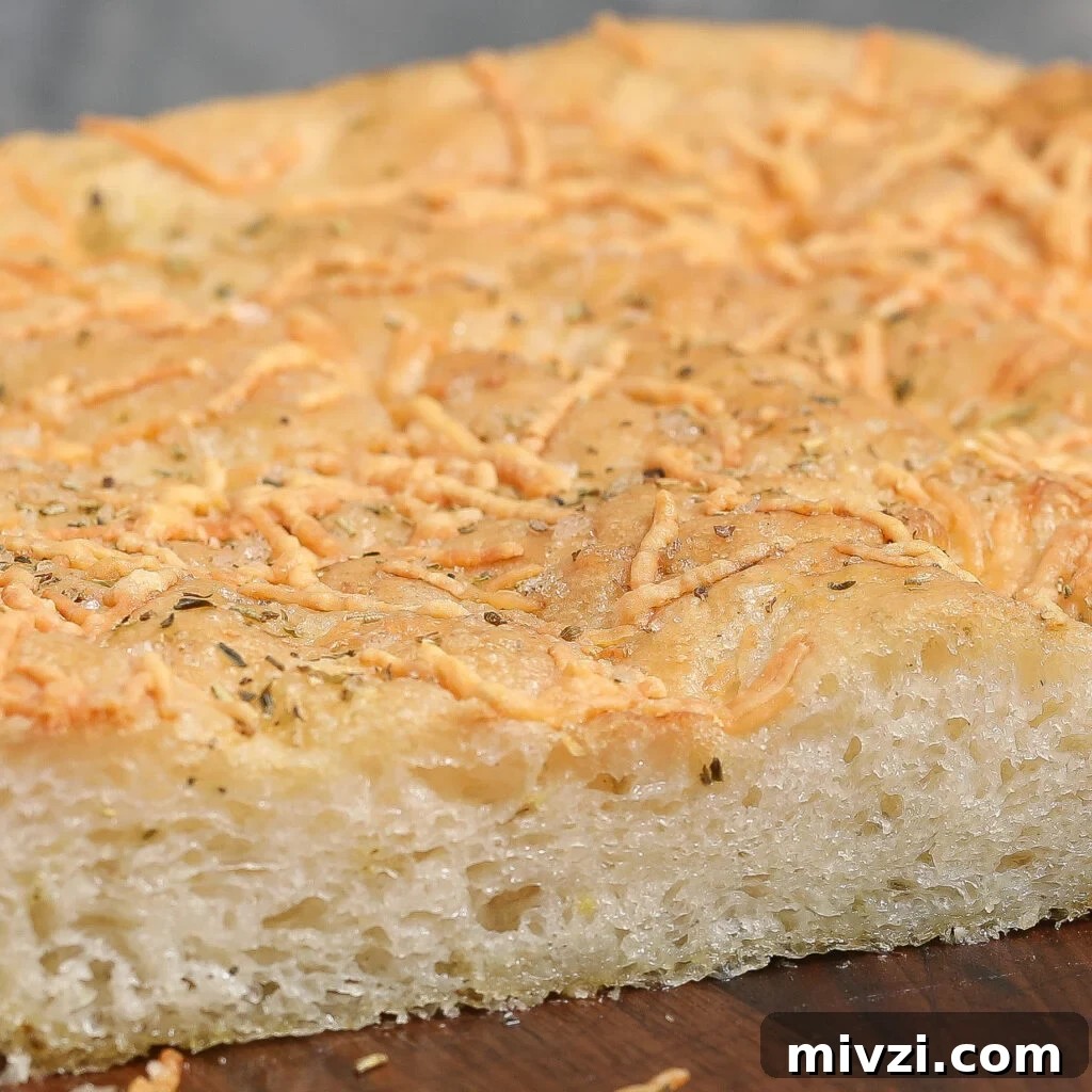 Parmesan and Herb Focaccia Recipe: Crispy, Fluffy Loaf 2 A close-up of a thick slice of Parmesan Herb Focaccia, topped with shredded cheese and herbs, showing a golden, slightly crisp crust and a soft, airy interior.