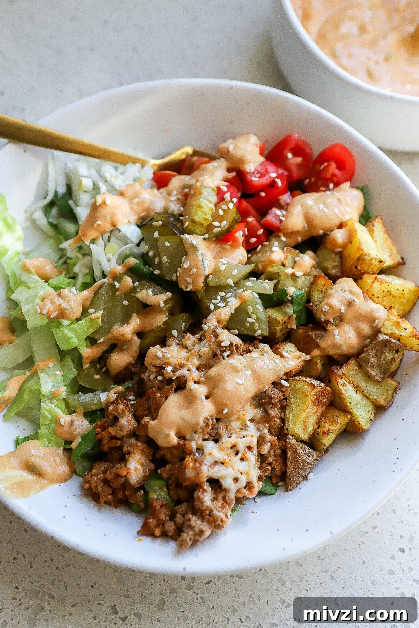 Side view of a loaded healthy burger bowl with a gold fork and special sauce in the background.
