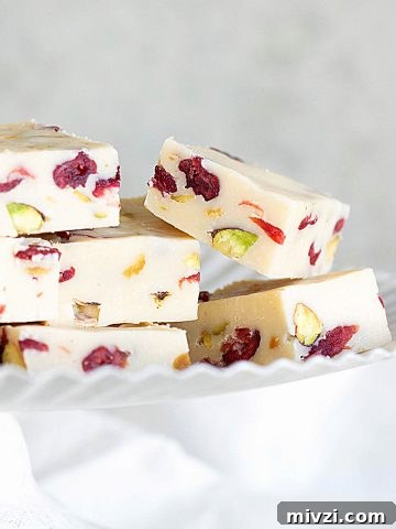 Several pieces of pistachio cranberry white chocolate fudge on a white cake stand with grey background.
