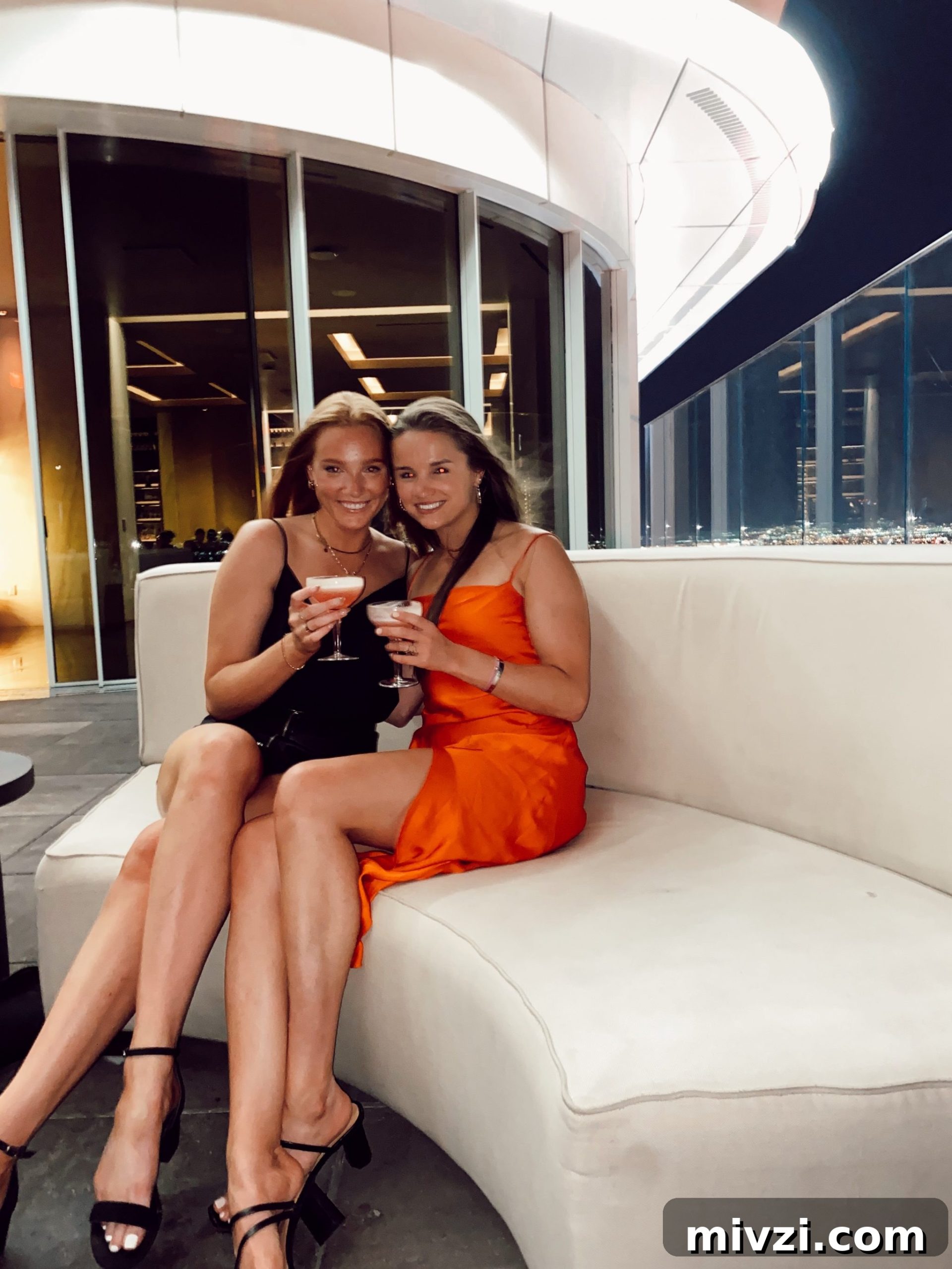 two girls in club attire sitting on white booth holding drinks overlooking skyline