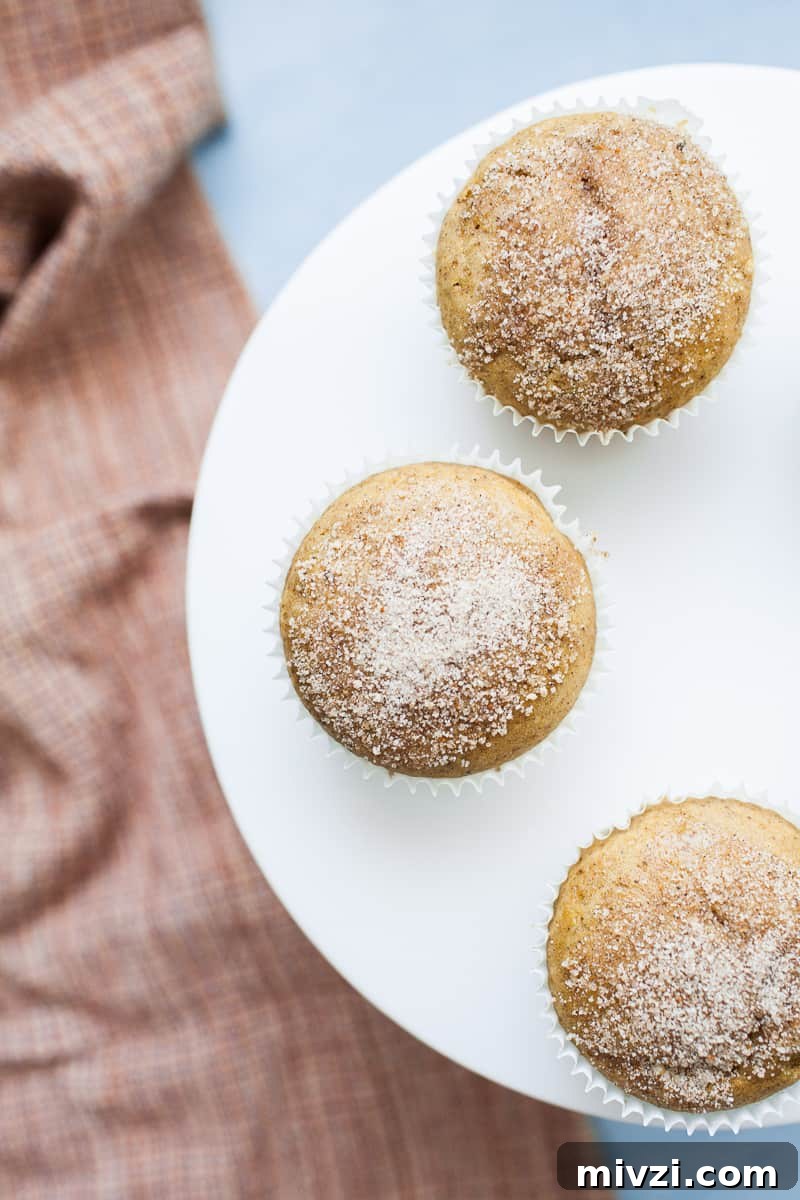 Three pumpkin muffins topped with cinnamon sugar sitting on top of a white cake stand with an orange plain napkin below.