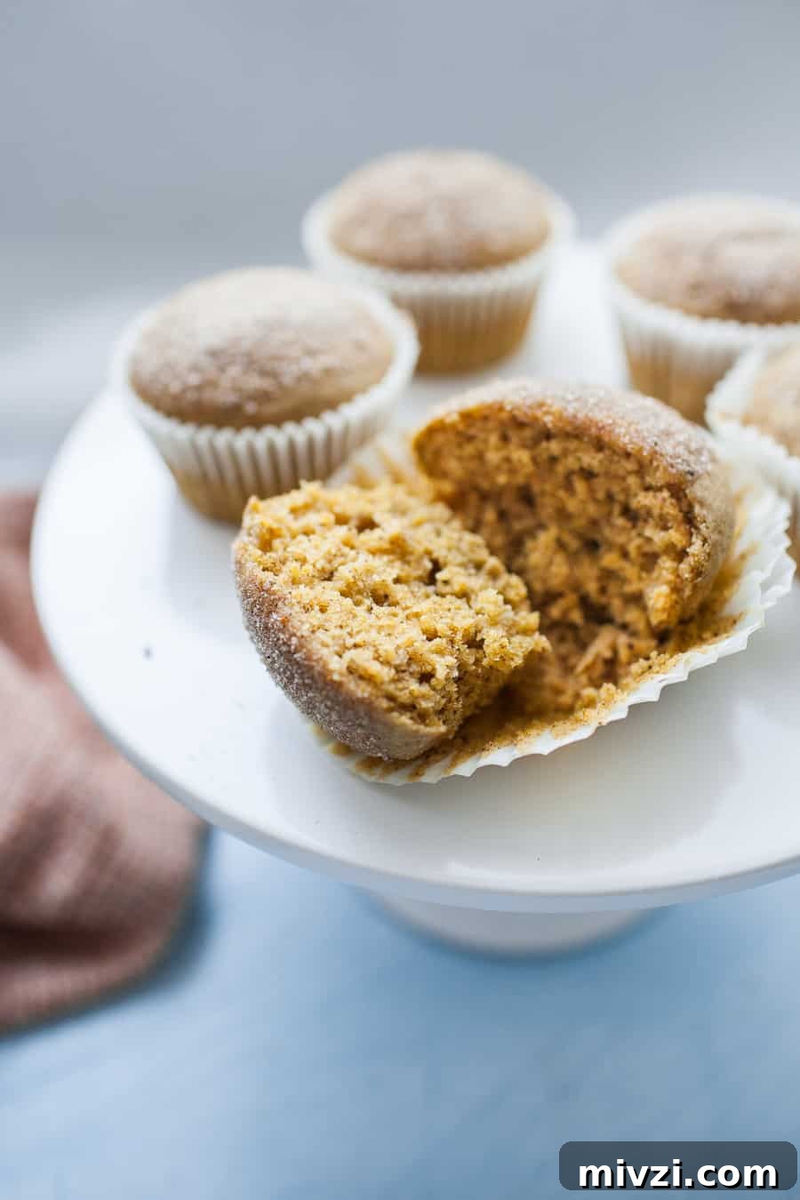 A pumpkin muffin, wrapped in a cupcake liner, cut in half sitting on top of a white porcelain cake stand.
