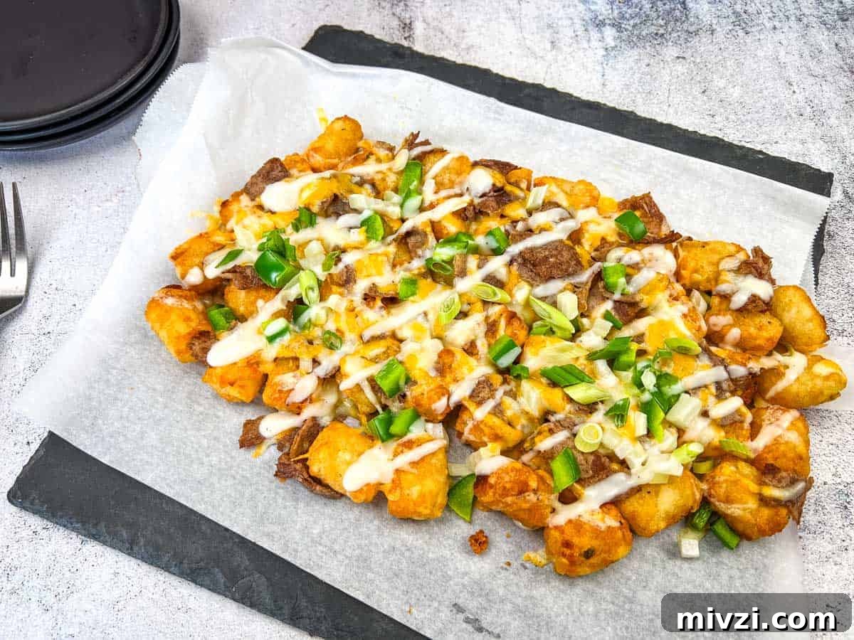 37 Winter Olympics Dishes to Warm Cold Nights and Long Broadcasts 36 Philly Cheesesteak Loaded Tots on parchment paper, topped with Alfredo cheese, green onions, and a drizzle of white sauce, placed on a dark rectangular slate plate.