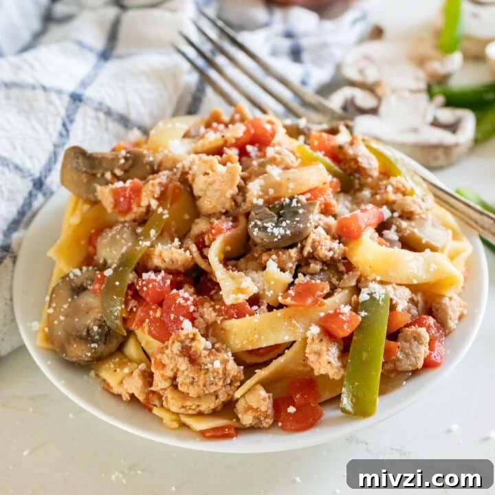 Sausage and peppers pasta on a white plate with a napkin, onion and mushrooms in the background and a fork across the back of the plate.
