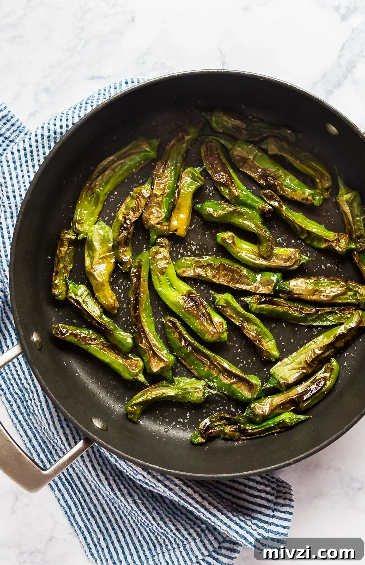 An overhead image of a pa with shishito peppers in it and a blue striped napkin.