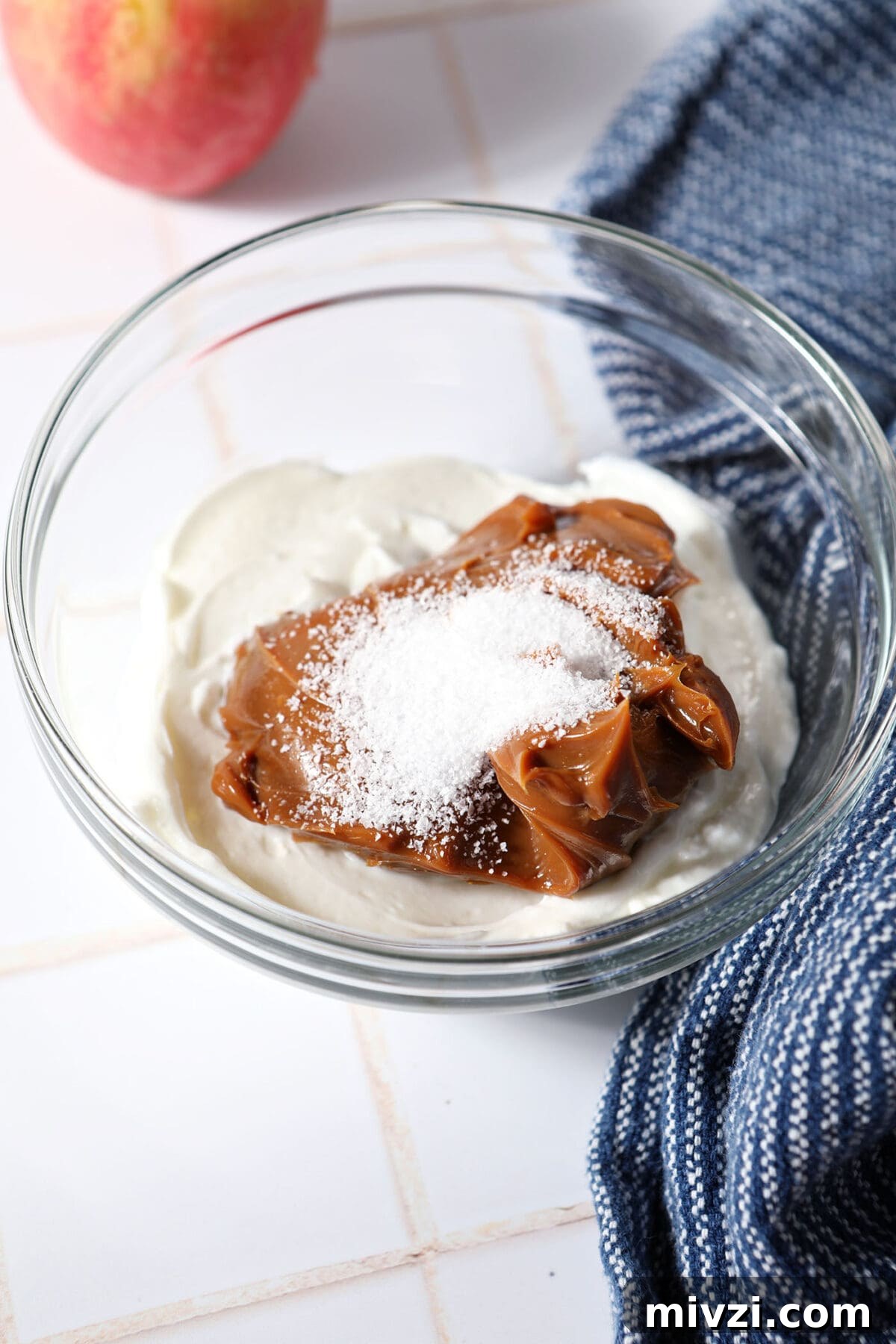 Creamy Dulce de Leche Dip Recipe for Desserts and Snacks 4 Greek yogurt, dulce de leche and salt in a mixing bowl on a white tiled surface next to a blue linen