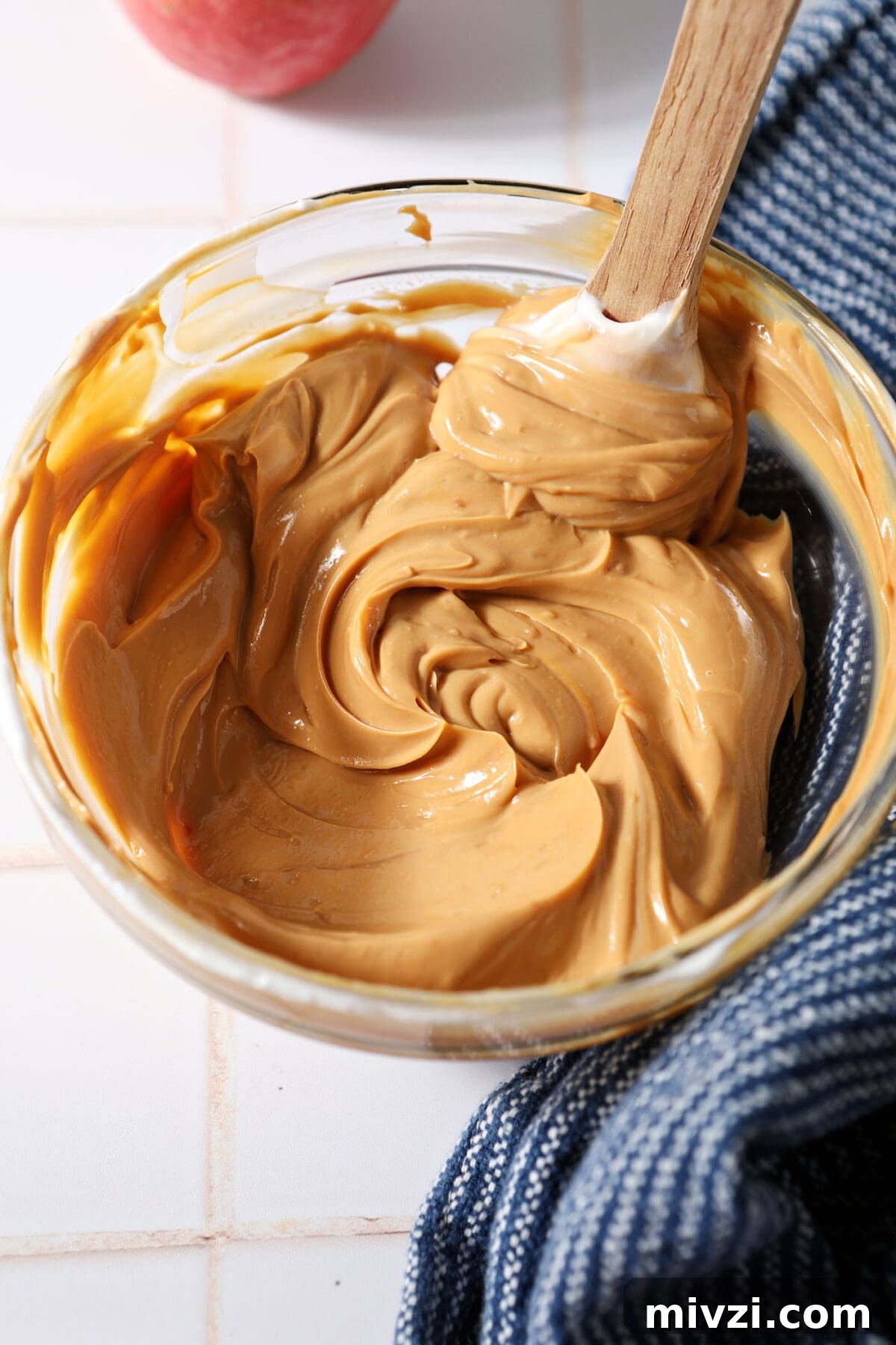 Creamy Dulce de Leche Dip Recipe for Desserts and Snacks 5 Mixed Dulce de Leche Caramel Dip in a bowl with a rubber spatula on a white tiled surface next to a blue linen