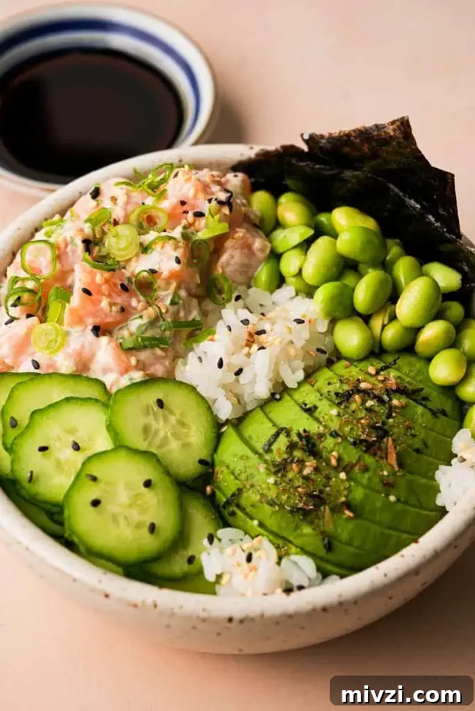 salmon poke bowl garnished with sliced avocado, cucumber, and edamame beside a small saucer of ponzu sauce