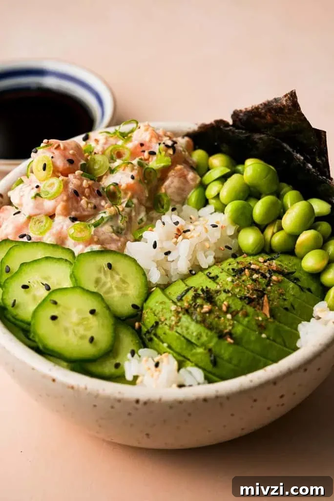 salmon poke bowl garnished with sliced avocado, cucumber, and edamame beside a small saucer of ponzu sauce