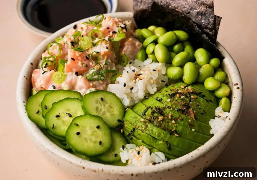 salmon poke bowl garnished with sliced avocado, cucumber, and edamame beside a small saucer of ponzu sauce