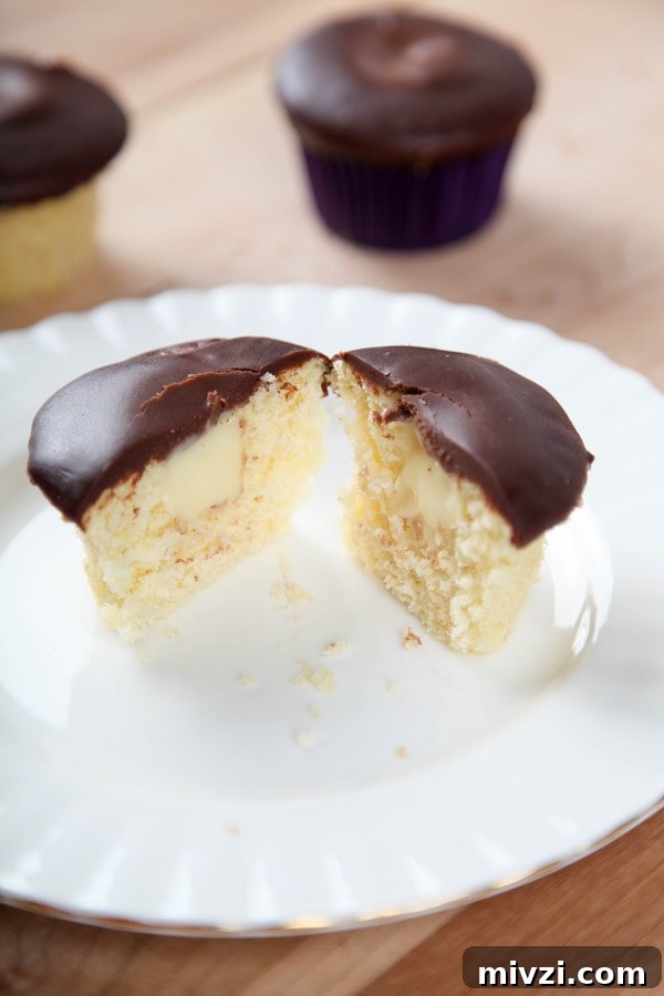 Boston Cream Pie Cupcakes Recipe with Vanilla Custard Filling 5 a cupcake topped with chocolate ganache cut in half to reveal a pastry cream center.