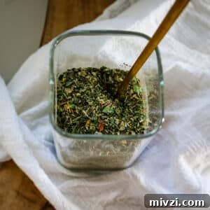 Italian seasoning in a clear jar with a spoon in it.