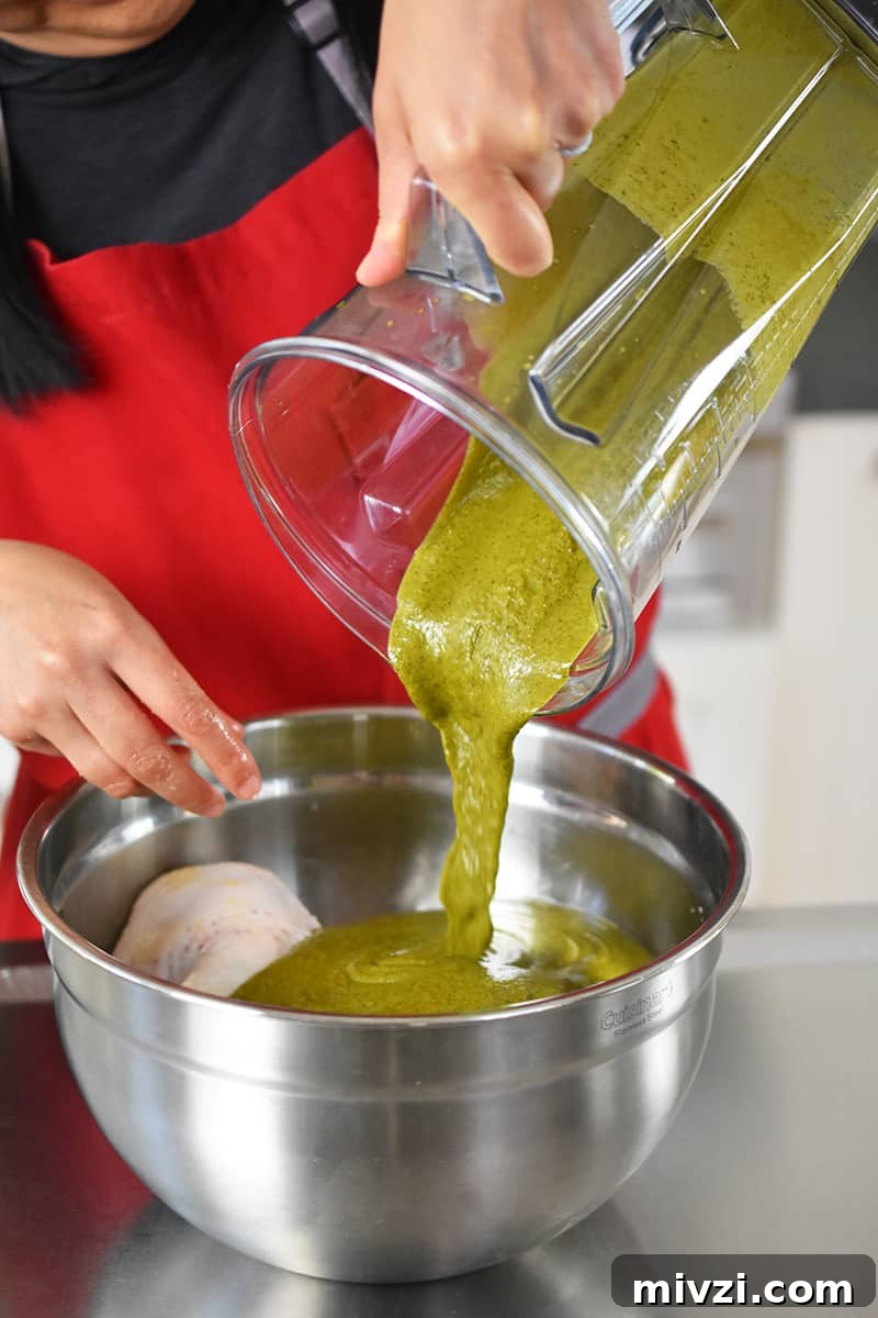 Gregory Gourdet’s Favorite Grilled Chicken Recipe 6 A hand is pouring a greenish brown marinade into a silver mixing bowl filled with salted chicken thighs.