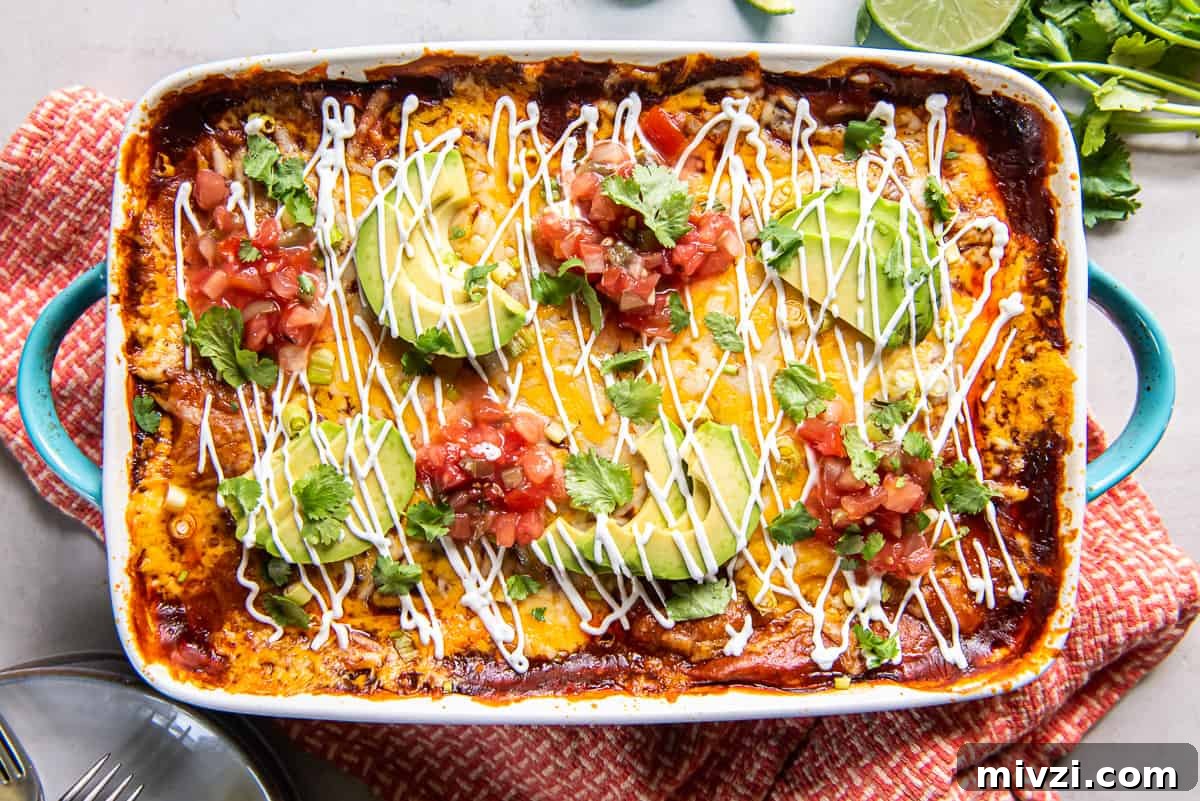 A top down shot of a baking dish filled with chicken enchilada casserole topped with sliced avocado and sour cream on an orange and white kitchen towel.