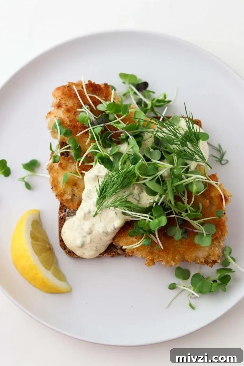 Pan-fried fish on rye bread topped with sauce, greens and with a lemon wedge.