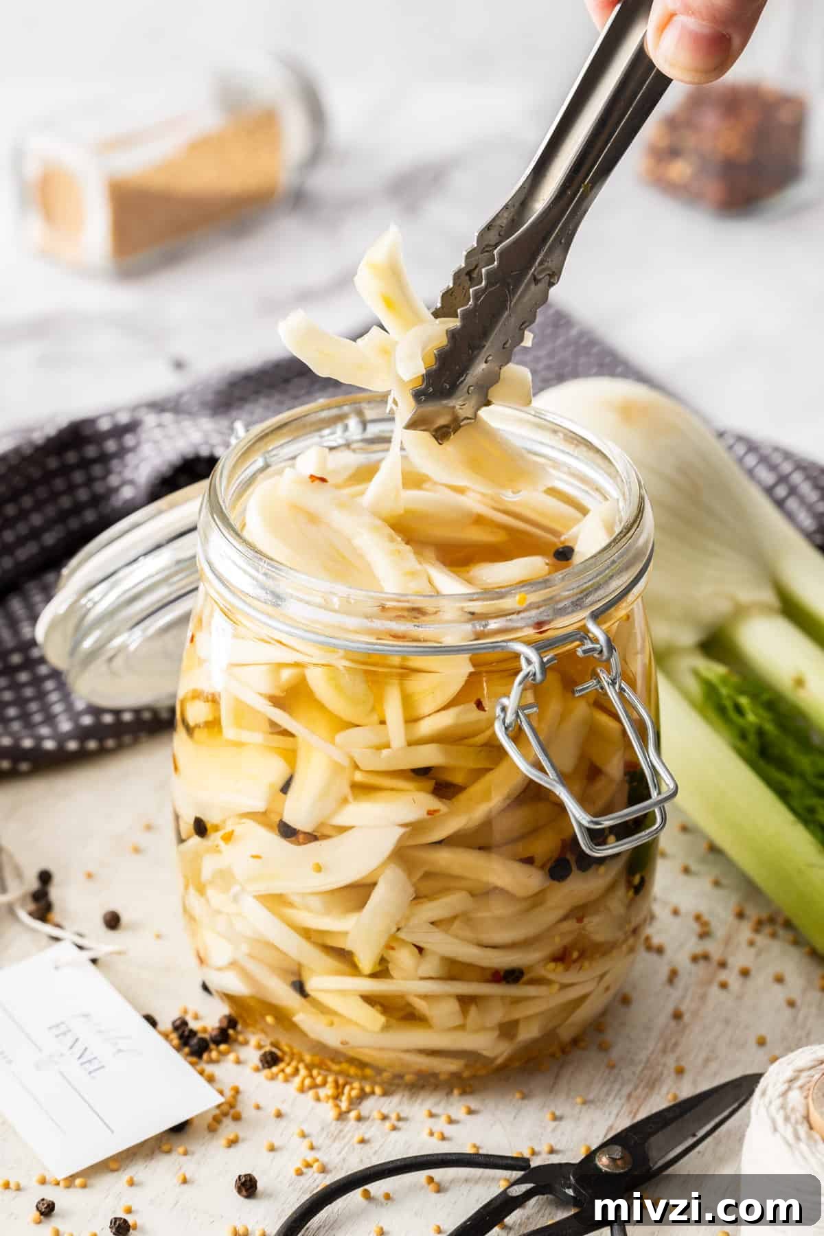 Jar of Pickled Fennel, sitting on a wooden board, with some tongs reaching in to pick up some pieces of fennel.