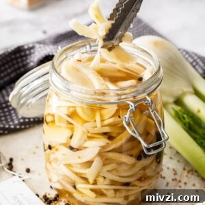 Jar of Pickled Fennel, sitting on a wooden board, with some tongs reaching in to pick up some pieces of fennel.