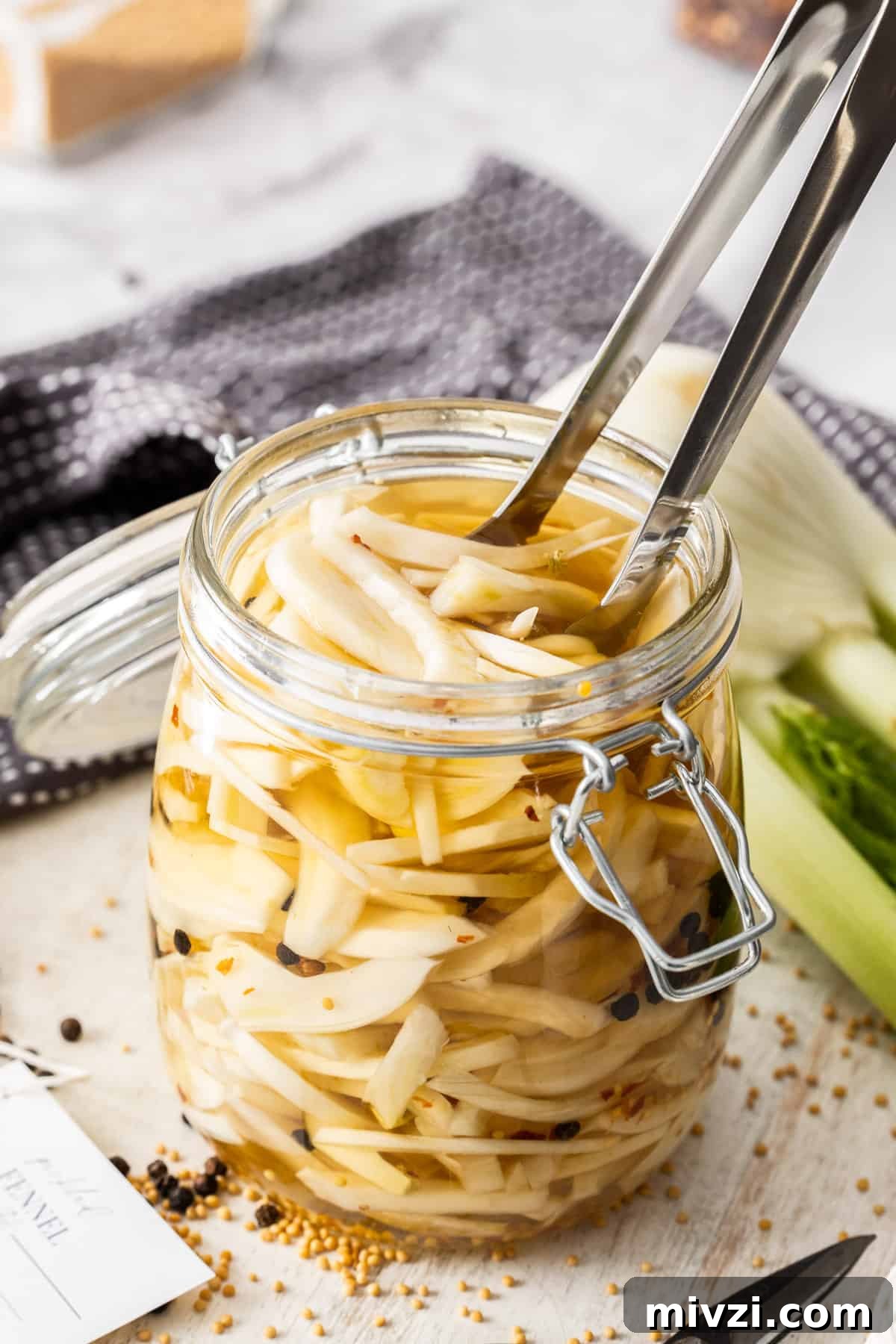 Jar of Pickled Fennel, sitting on a wooden board, with some tongs reaching in to pick up some pieces of fennel.