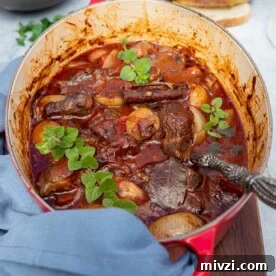A Greek beef stew or stifado in a red cast iron pot on a wooden board with a blue tea towel and with bread and bowls in the background.