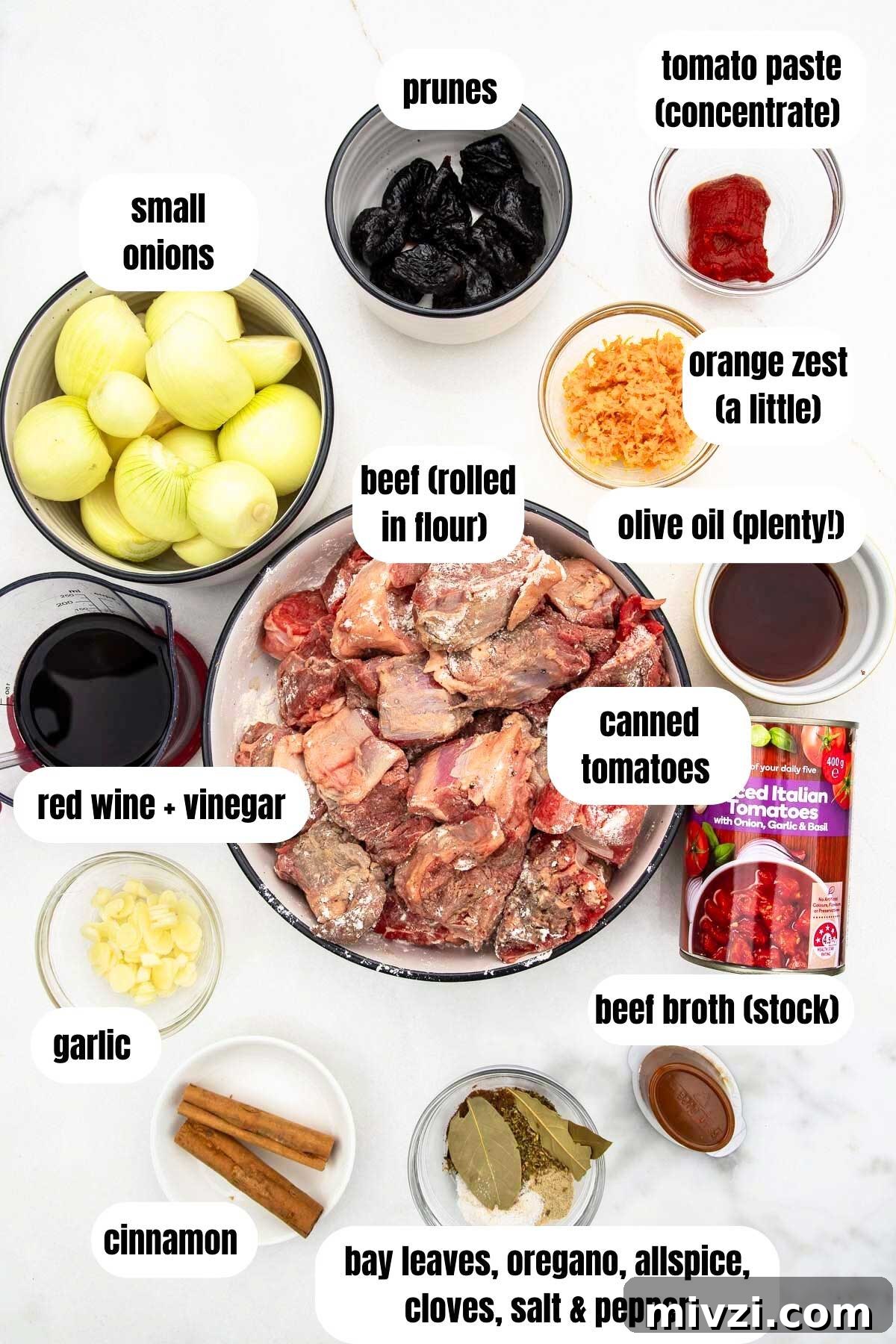 An overhead view of all the ingredients needed to make a Greek beef stew including stewing beef, bay leaves, oregano, allspice, cloves and salt and pepper, beef broth, cinnamon sticks, chopped garlic, canned tomatoes, red wine and vinegar, olive oil, orange zest, tomato paste, prune and small onions.