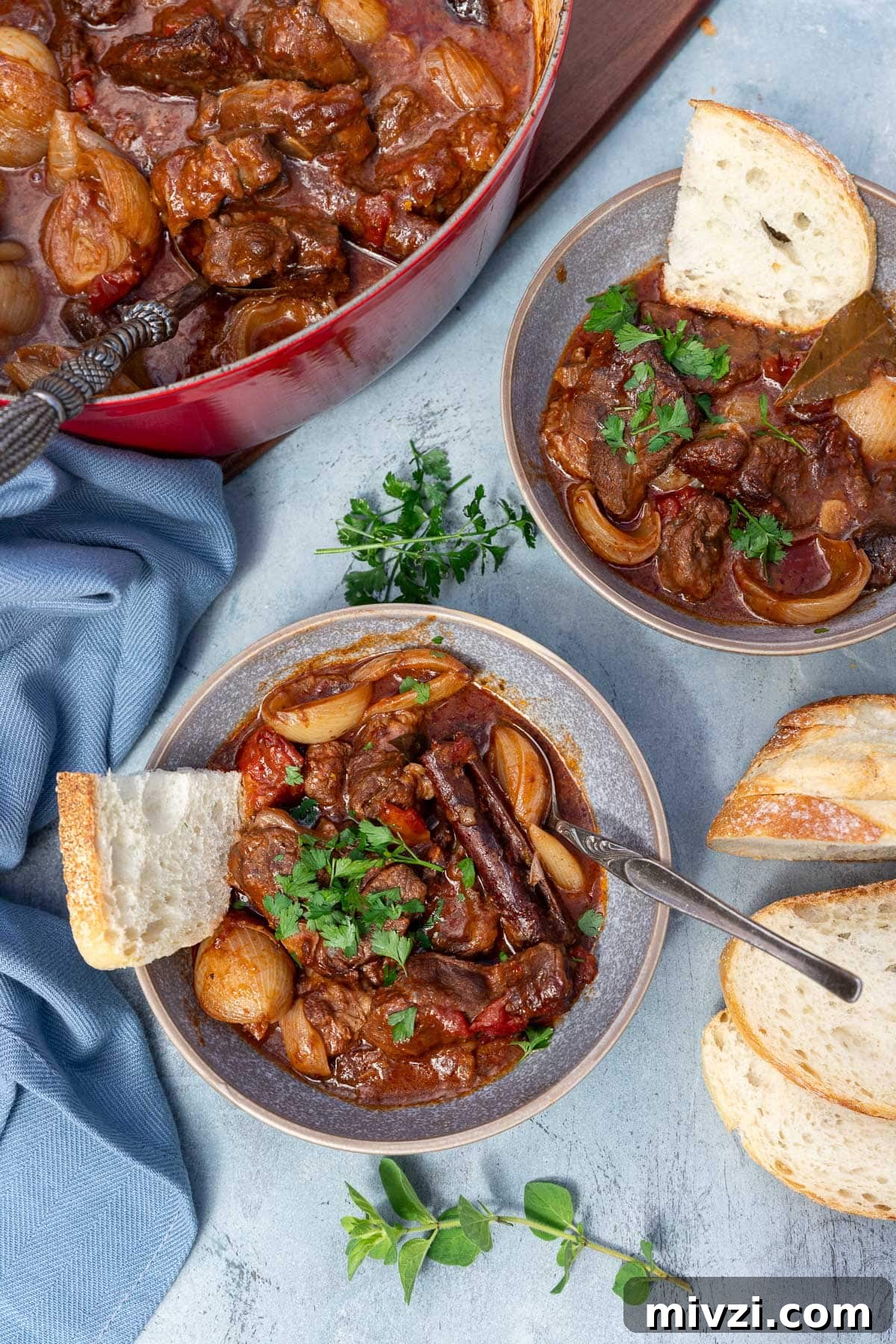 Overhead view of two bowls of Greek beef stew with spoon and a piece of bread in it with bread and a pot of stew behind, on a blue background.