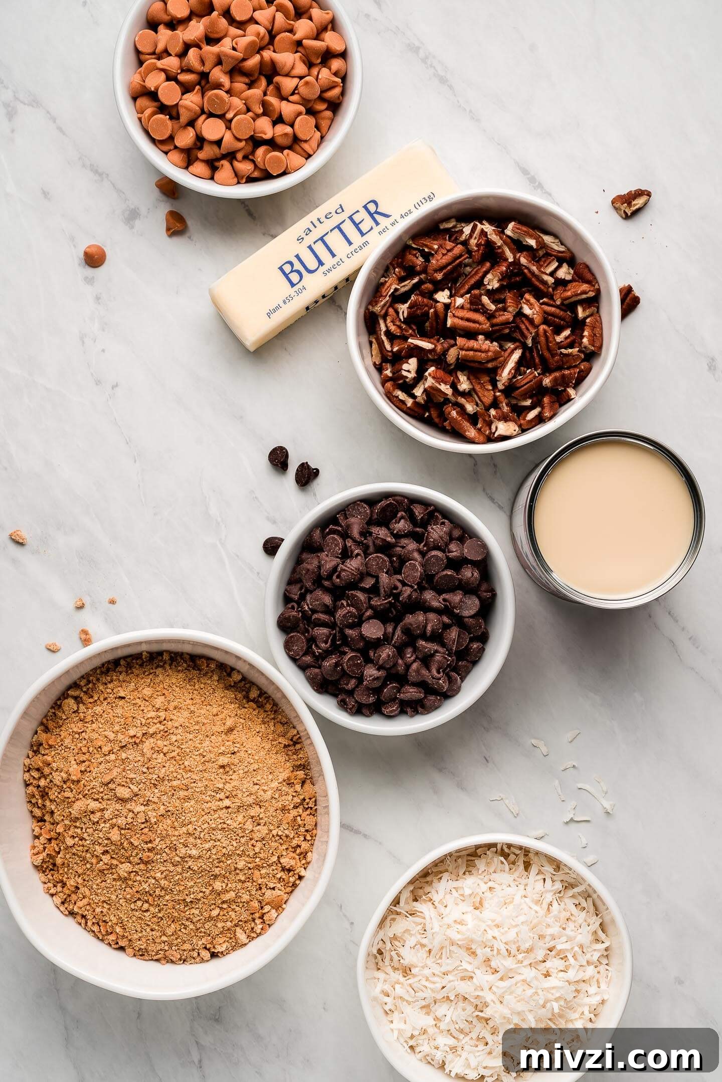 Ingredients on a marble surface- butterscotch chips, pecans, condensed milk, chocolate, graham cracker crumbs, and coconut.