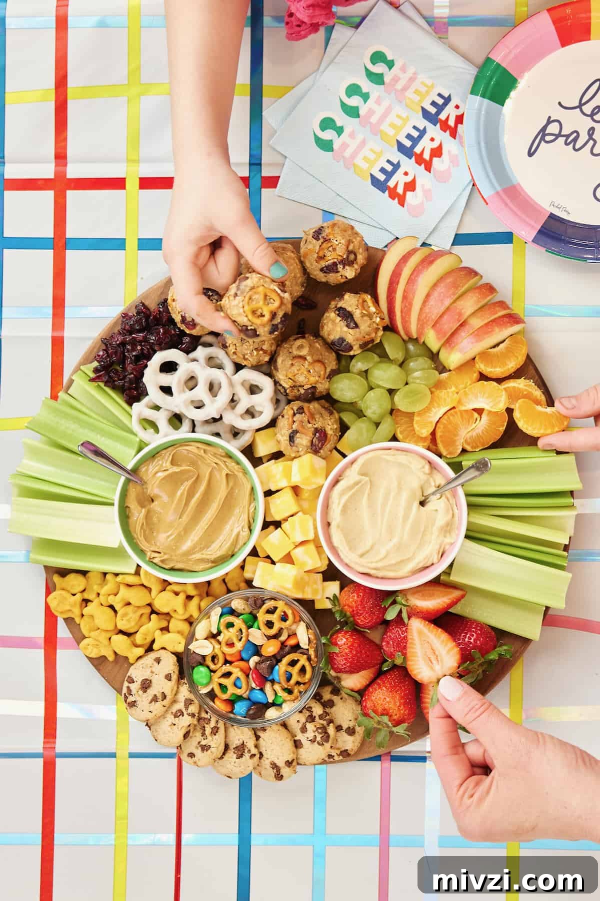 A round wood board with hummus, peanut butter, snack mix, cheese cubes, apple slices, mandarin orange segments, grapes, strawberries, Goldfish crackers, and mini cookies.