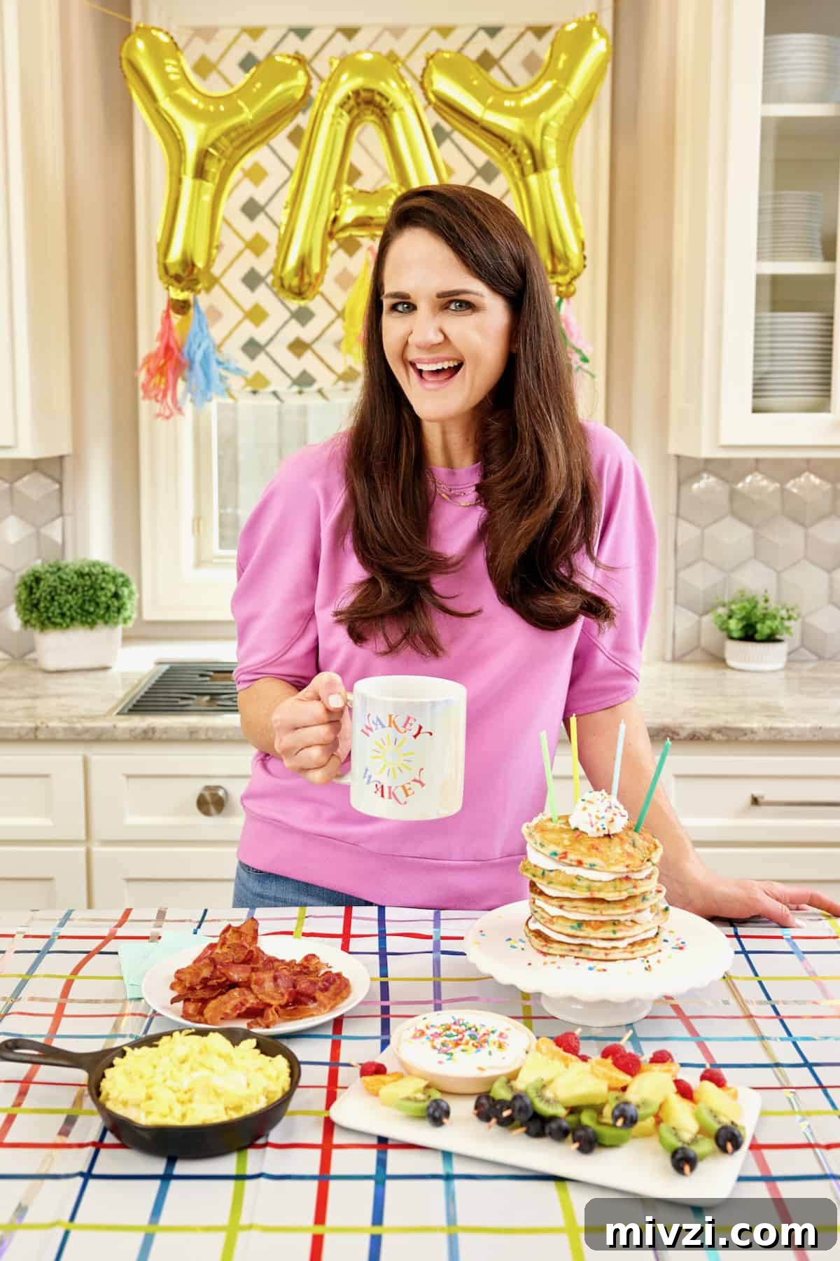 A breakfast spread featuring soft scrambled eggs in black cast iron skillet, bacon on a round white plate, a stack of confetti pancakes on a white cake plate, and a pile of fruit skewers.