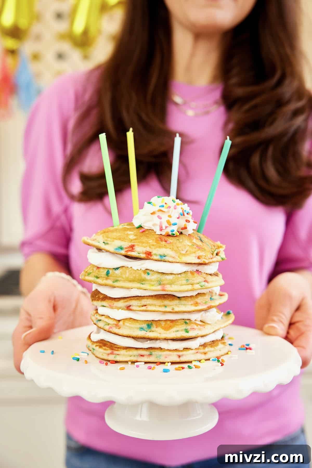 A stack of five confetti pancakes with layers of whipped topping between on a white cake plate with four candles on top.