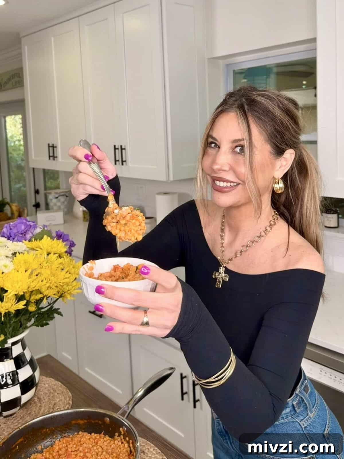 A smiling woman with long brown hair holds a bowl of food in one hand and a spoonful in the other, standing in a bright kitchen with white cabinets and a vase of yellow and purple flowers on the counter.