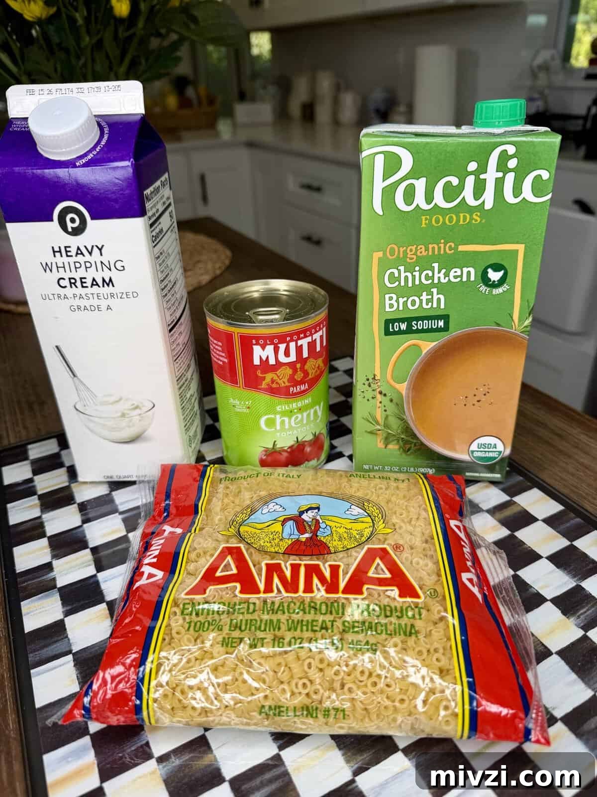 A carton of heavy whipping cream, a carton of Pacific Foods organic chicken broth, a can of Mutti cherry tomatoes, and a bag of Anna ditalini pasta are arranged on a checkered countertop in a kitchen.