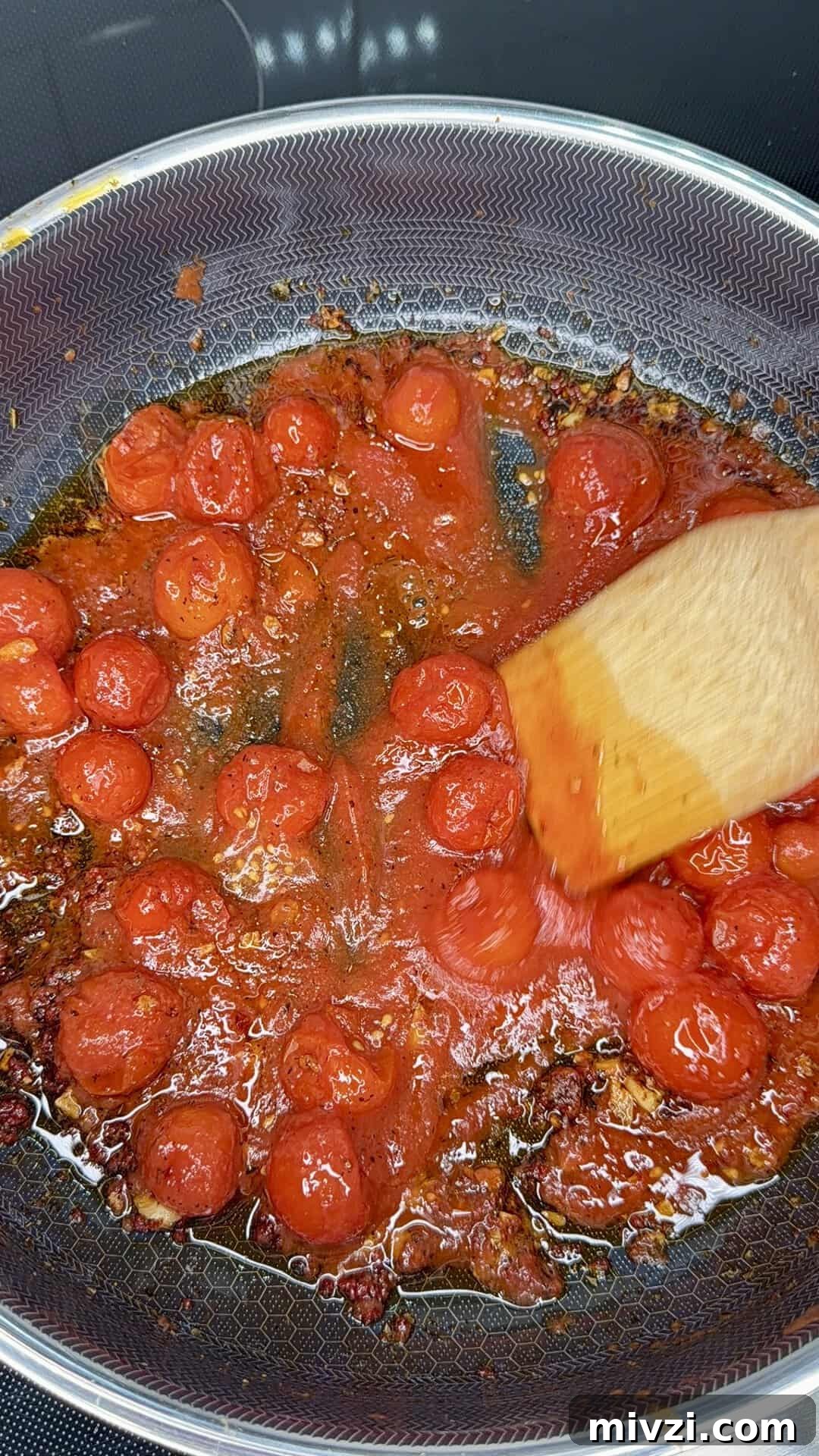 Cherry tomatoes cooking in a nonstick pan with oil and spices, being stirred with a wooden spatula; the tomatoes are beginning to break down and release their juices.