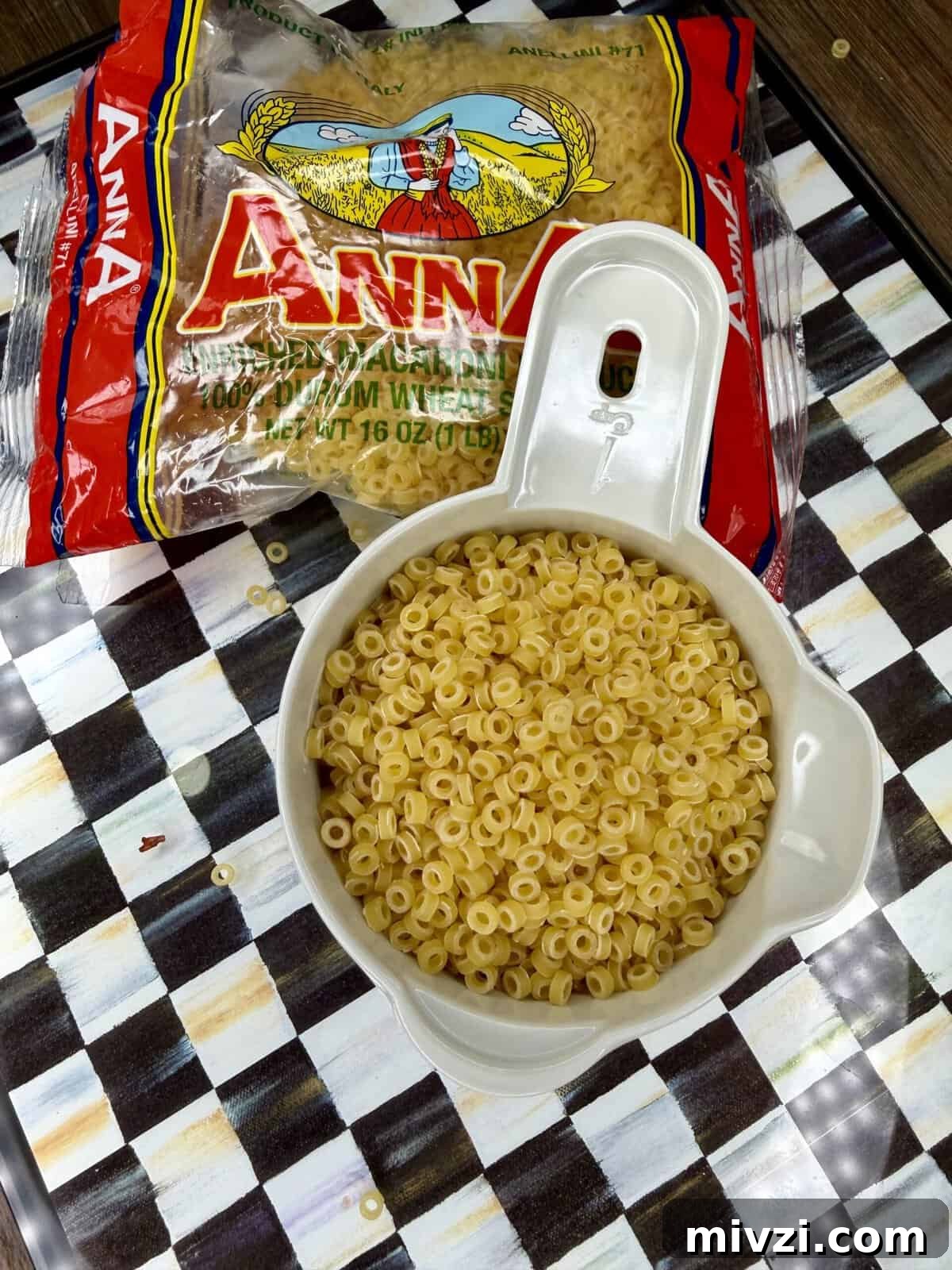A measuring cup filled with dry ditalini pasta sits on a checkered surface, with a partially opened bag of Anna brand ditalini pasta in the background. Some pasta pieces are scattered around.