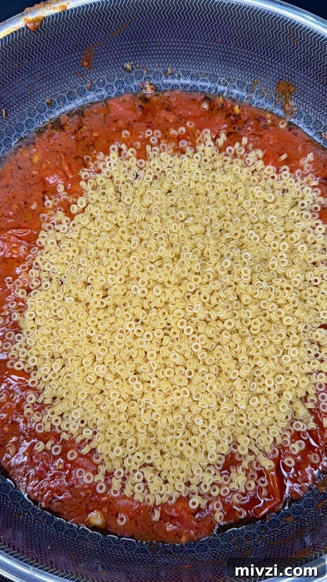 A close-up of a pan with tomato sauce and a large pile of dry ring-shaped pasta sitting on top, not yet mixed together.