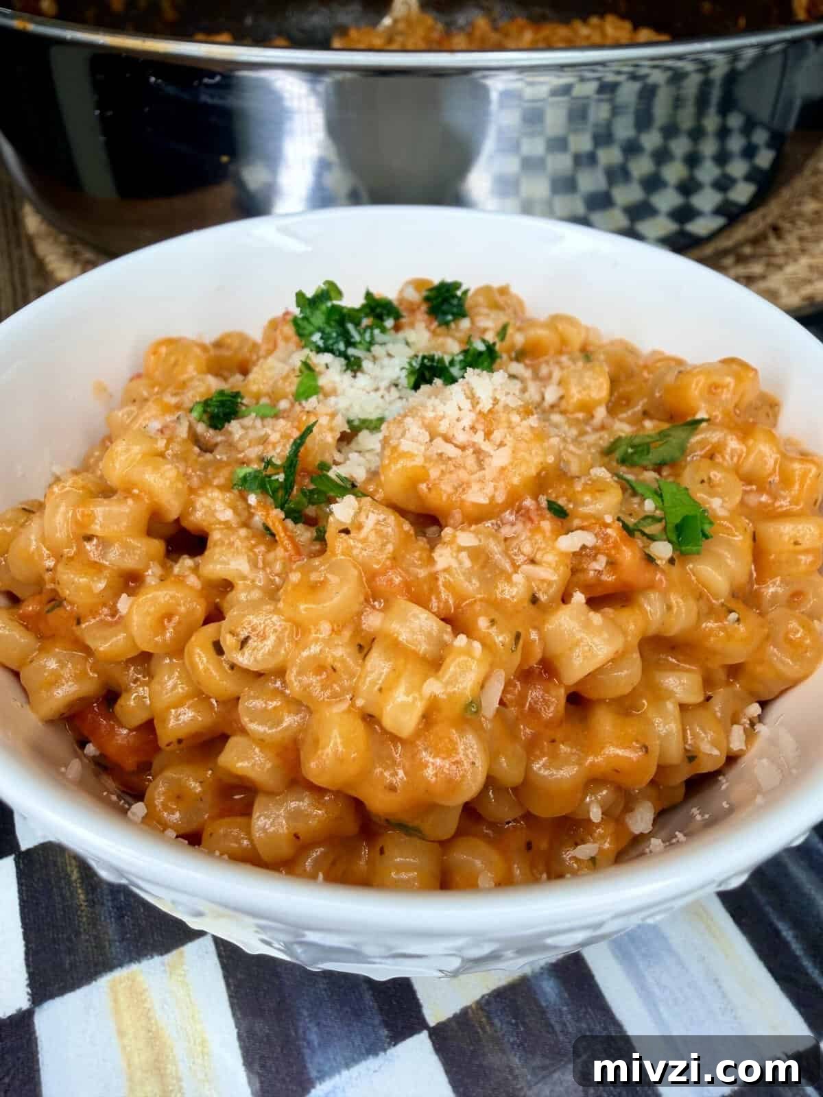 A bowl of creamy pasta with small tube-shaped noodles in a rich orange sauce, topped with grated cheese and fresh parsley. A pan of more pasta is visible in the background.
