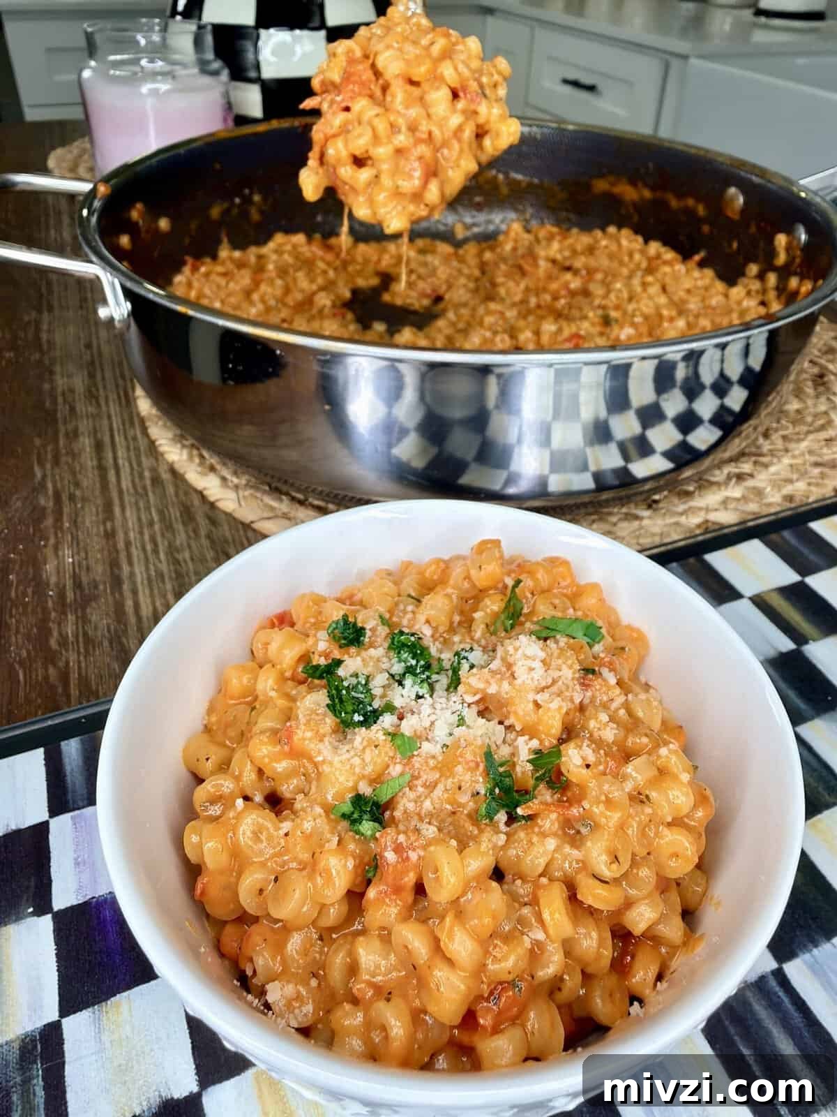 A bowl of creamy macaroni pasta topped with herbs and breadcrumbs sits on a table, with a large pan of the same pasta in the background and a serving spoon lifting a portion from the pan.