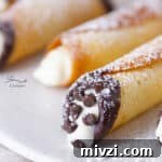 close up on the ends of cannoli cookies, the main focus is a chocolate dipped end sprinkled with chocolate chips, all are dusted with powdered sugar.