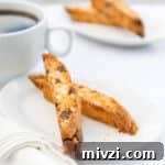 two pieces of biscotti on a white plate with a white background and a cup of coffee, more biscotti in the back.