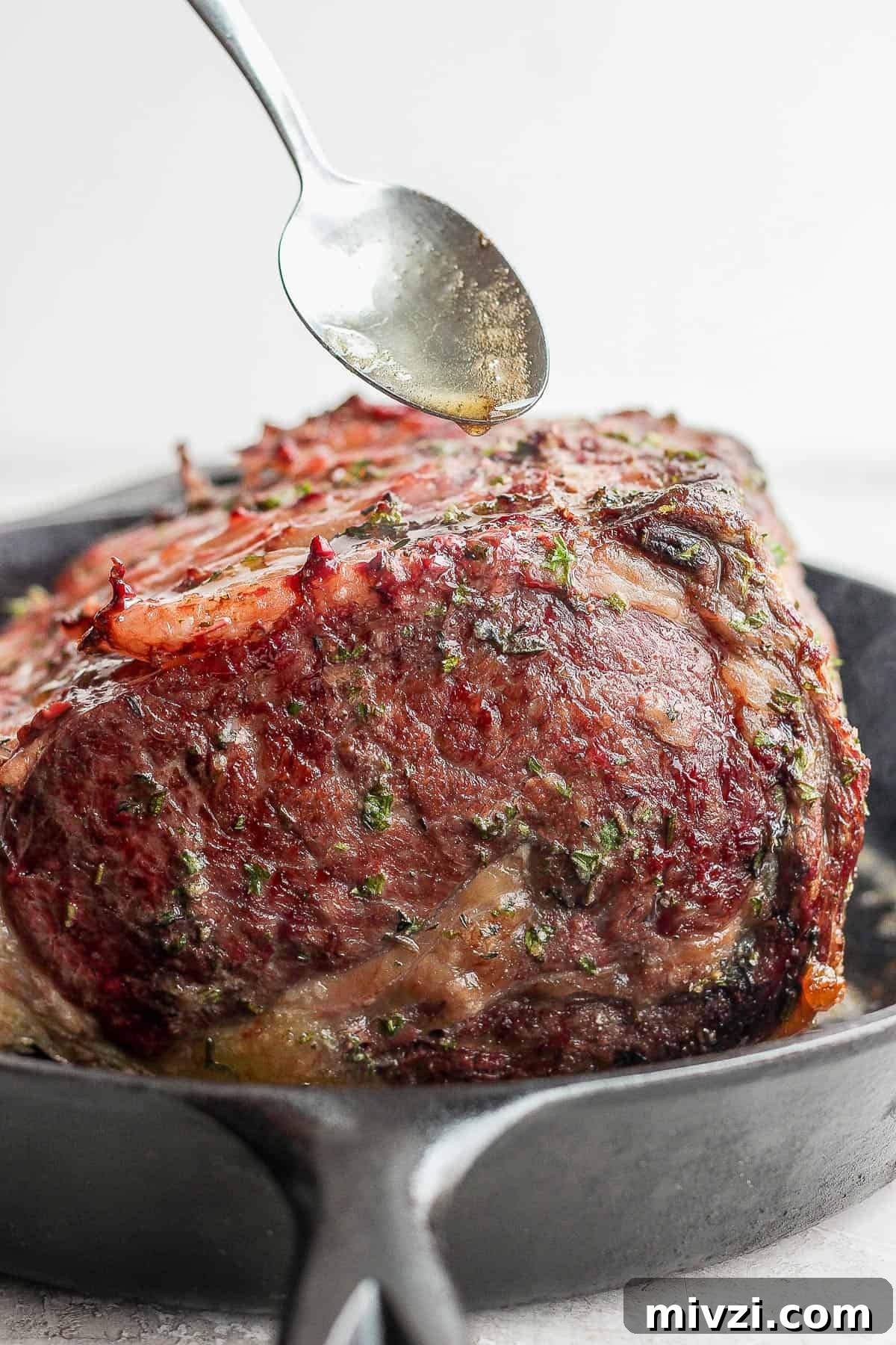 Prime rib being seared in a cast iron skillet and a spoon scooping the rendered fat over the top.