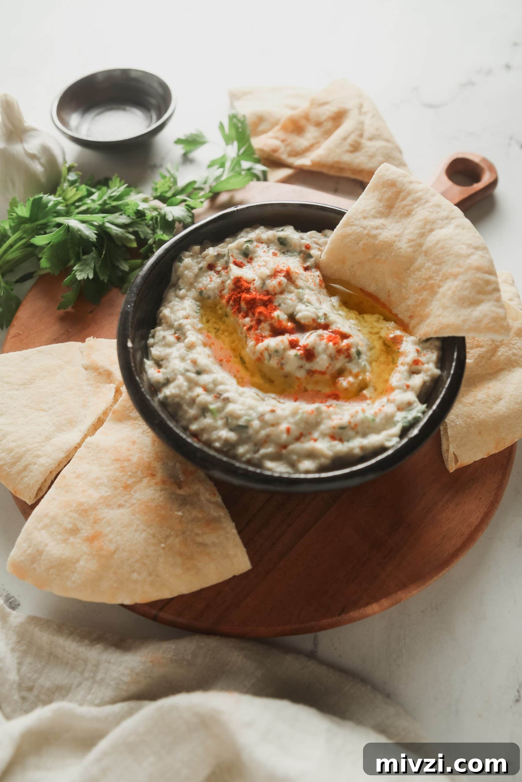 A cutting board with a bowl of baba ganoush and pita wedges.