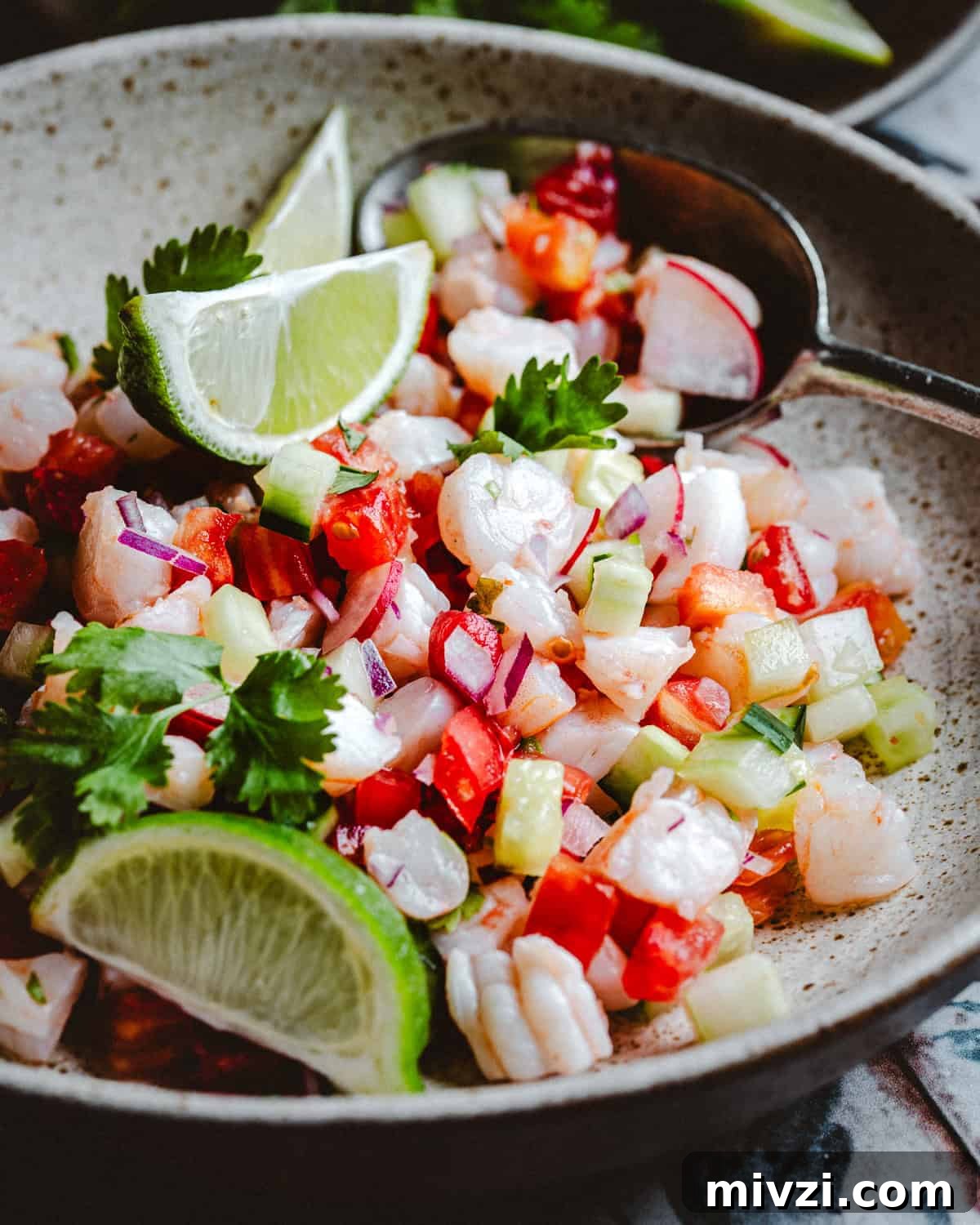 A close-up of a bowl of shrimp ceviche with diced tomatoes, red onions, cucumbers, cilantro, and radishes, garnished with lime wedges and served with a spoon.