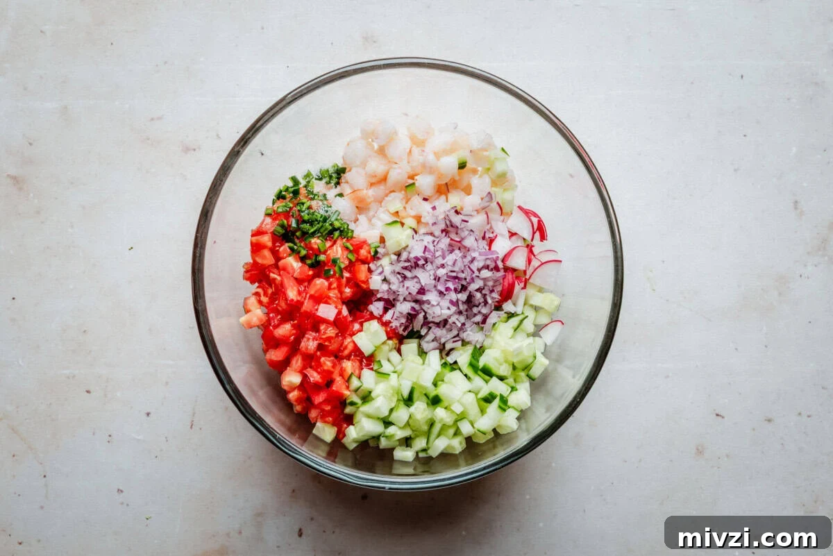 A glass bowl containing neatly arranged diced shrimp, tomato, cucumber, red onion, radish, and chopped herbs on a light surface.