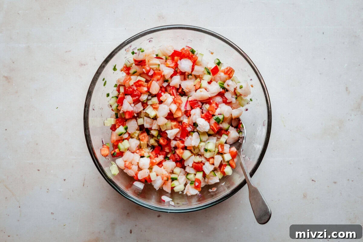 A glass bowl filled with diced shrimp, tomatoes, cucumbers, onions, and herbs, with a spoon resting inside, sits on a light-colored surface.