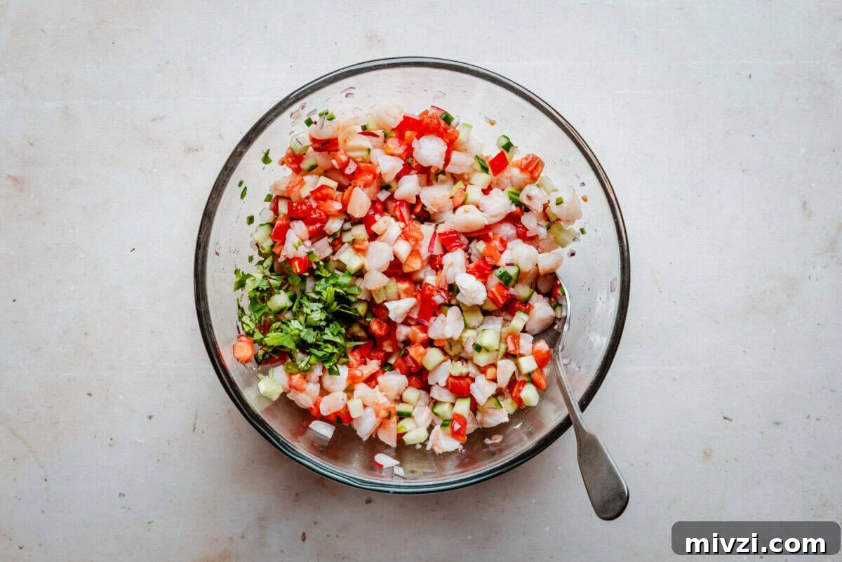 A glass bowl filled with ceviche made of diced shrimp, tomatoes, onions, cucumbers, and cilantro, with a spoon resting inside. The bowl sits on a light-colored surface.