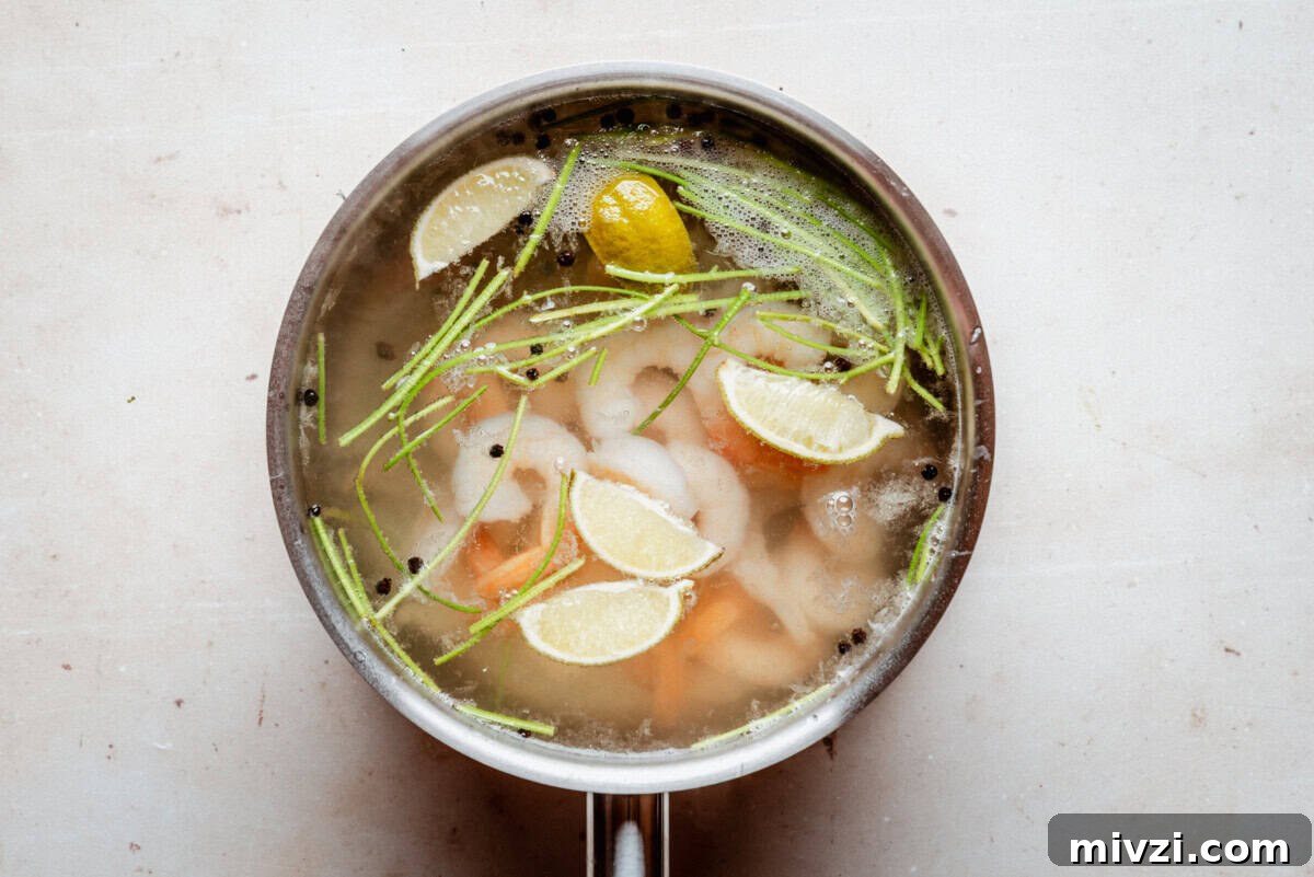 A pot of broth with shrimp, lemon wedges, green herbs, and black peppercorns floating on the surface, viewed from above on a light background.