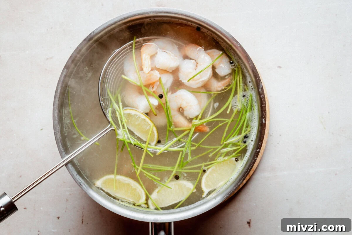 A pot of clear soup with shrimp, lime wedges, green herbs, and black peppercorns. A slotted spoon is lifting some shrimp from the broth. The pot is on a light-colored surface.