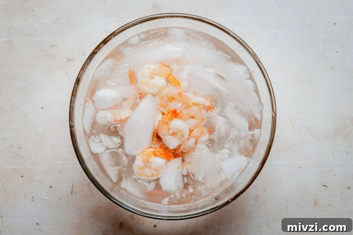 A glass bowl filled with shrimp and ice sits on a light-colored surface, with the shrimp partially submerged in water and surrounded by ice cubes.