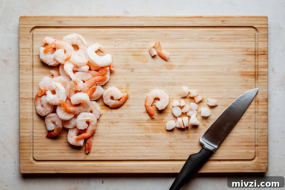 A wooden cutting board with shrimp—some whole, some chopped—next to a small kitchen knife with a black handle. The board is placed on a light-colored surface.
