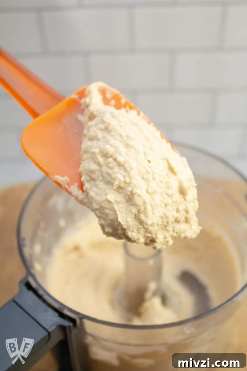 Classic Hummus from Canned Chickpeas – Quick Authentic Recipe 6 Scooping freshly made hummus from a food processor with a spatula.