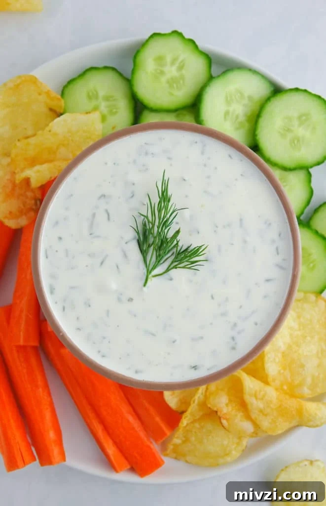 Greek yogurt dill pickle dip presented in a brown bowl on a plate with chopped veggies and potato chips. Food is on a white marble countertop.